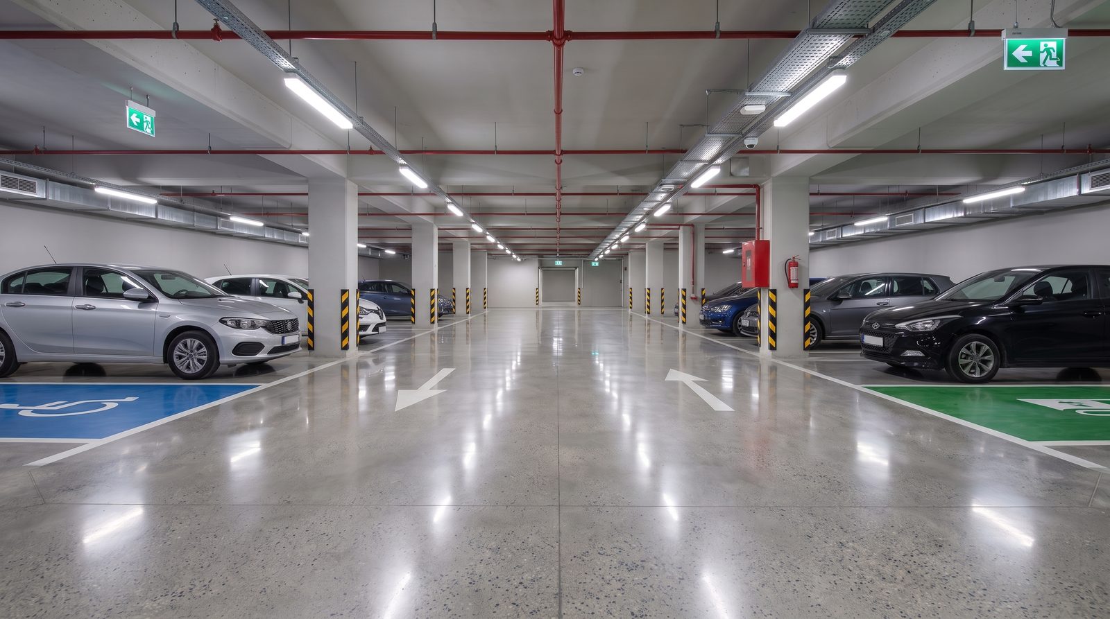 Parlak betonla kaplanmış kapalı katlı otopark katı; beyaz park çizgileri, sarı-siyah kolonlar ve tavan LED sıraları zeminde net yansıyor.