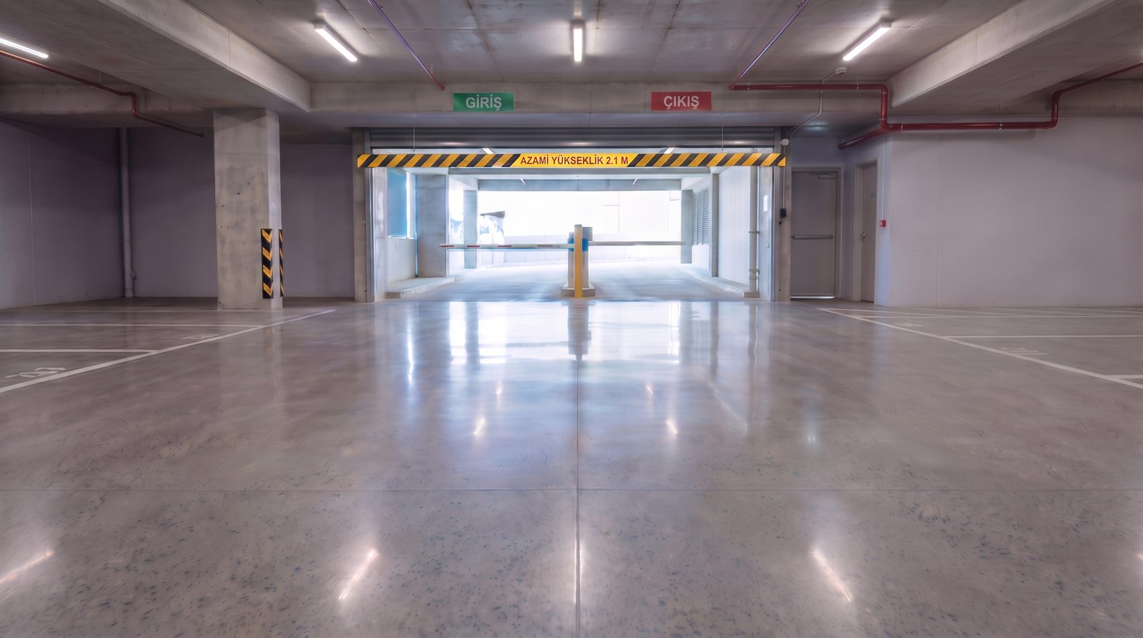 Kapalı otopark girişine yakın parlak beton kat platformu; sarı-siyah yükseklik levhası ve gün ışığı gelen giriş portalı görünüyor.